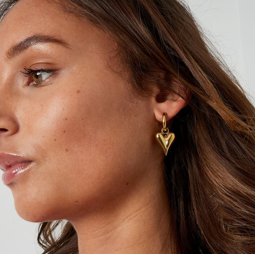 Close-up of a woman wearing gold heart-shaped earrings and necklace against a neutral background